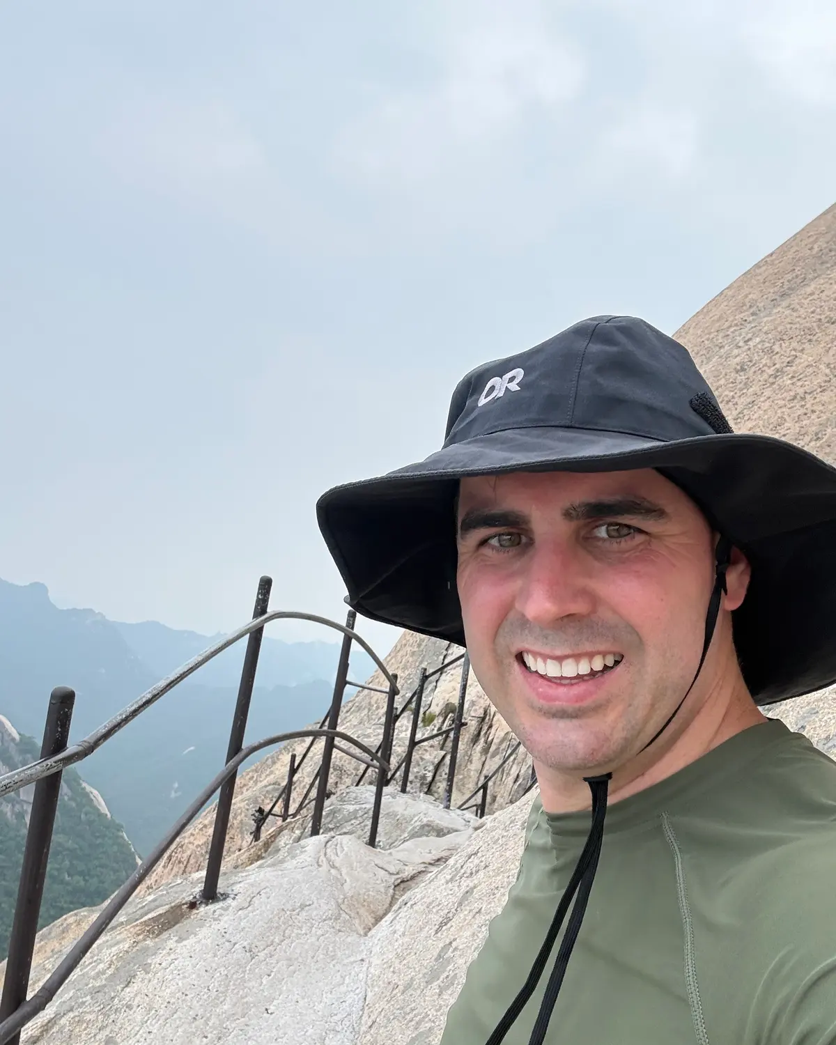 Nathan Brennan in a wide-brimmed sun hat on a rocky mountain trail, with a railing and hazy ridgelines behind him